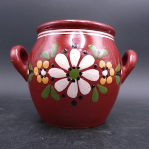 May include: A red ceramic pot with two handles and a floral design. The design features white flowers with yellow centers and green leaves.