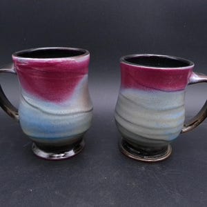 Puede incluir: Dos tazas de cerámica con un borde negro y un esmalte que es una mezcla de rojo, azul y gris. Las tazas tienen un cuerpo redondeado y un asa.