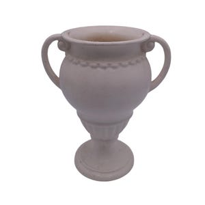Może przedstawiać: Biały ceramiczny wazon z dwoma uchwytami i podstawą na cokole. Wazon ma ozdobny wzór wokół górnej części.