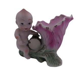 Puede incluir: Una figura de porcelana vintage que representa una figura de querubín con una cabeza rosada y un pequeño pañal, sosteniendo un objeto redondo. La figura está junto a un gran jarrón en forma de flor rosa con hojas verdes.