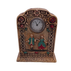 May include: A decorative clock with a carved wooden frame and a white face. The clock features a painted scene of two people dancing in front of a building with a floral design. The clock face has black Roman numeral hour markers and black hands.