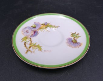 Peut inclure: Une soucoupe blanche avec un bord vert et or. La soucoupe présente un motif peint de chardons violets et verts avec une signature qui se lit "E. Garnier".