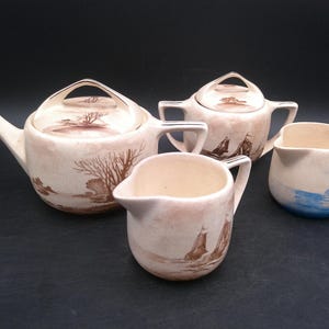 Può includere: Un servizio da tè vintage con teiera, zuccheriera, lattiera e tazza. Il set presenta uno sfondo color crema con disegni di paesaggi e barche a vela marroni. La tazza ha un disegno dipinto di blu.