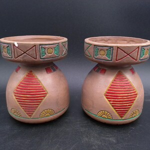 May include: Two brown ceramic candle holders with geometric designs in red, green, and yellow. The holders have a wide base and a narrow top.