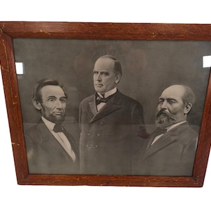May include: A framed black and white photograph of three men in formal attire. The men are standing in a row, with the man in the middle wearing a dark suit and a bow tie. The men on either side are wearing suits and ties. The photograph is in a wooden frame.