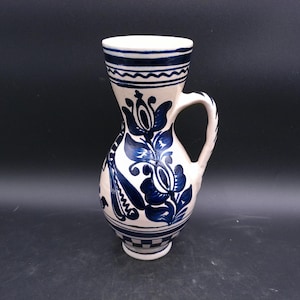 May include: A white ceramic pitcher with a blue floral design. The pitcher has a handle and a wide mouth.