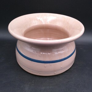 Puede incluir: Una maceta de cerámica con un borde ancho y acampanado y un cuerpo redondeado. La maceta es de color rosa pálido con una sola franja azul oscuro alrededor del centro. El interior es de un tono rosa más oscuro. La maceta es probablemente para uso en interiores o exteriores.