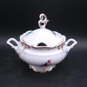 Puede incluir: Sopera de porcelana blanca con tapa y asas. Decorada con motivos florales coloridos y ribetes dorados. La tapa tiene un asa decorativa y una abertura para la cuchara. Una pieza clásica para servir sopa o guiso.