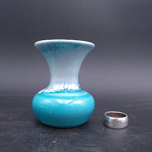 Könnte beinhalten: Eine kleine, türkisfarbene Keramikvase mit weißer, getropfter Glasur im oberen Bereich. Rechts von der Vase liegt ein Silberring. Die Vase hat einen abgerundeten Boden und eine ausgestellte Öffnung. Der Ring ist ein schlichtes Band.