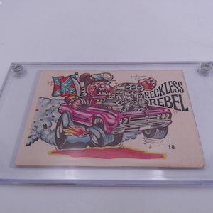 Puede incluir: Una ilustración de dibujos animados de un coche rosa con un conductor que lleva un sombrero y una bandera confederada en el fondo. El coche tiene un motor grande y el texto "Reckless Rebel" está impreso en el lateral. El número 18 está impreso en la esquina inferior derecha.