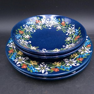 May include: A set of three blue ceramic plates with a white floral design. The plates feature a white flower with a yellow center and green leaves.