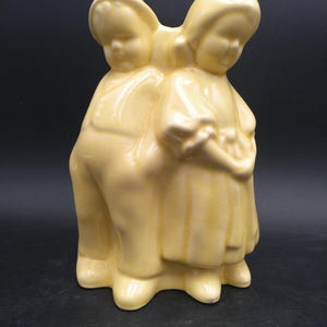 Könnte beinhalten: Eine Vintage-Keramikfigur, die zwei Kinder darstellt, einen Jungen und ein Mädchen, die eng beieinander stehen. Die Figur ist in einem weichen Gelb gehalten und hat eine glänzende Oberfläche. Der Junge trägt eine Latzhose, und das Mädchen trägt ein Kleid.