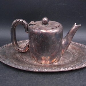 May include: Vintage silver-plated teapot and oval tray. The teapot has a curved handle with decorative perforations and a small spout. The tray has an ornate, embossed border. The teapot has the initials "H.E." engraved on the side.