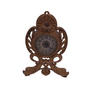 May include: A gold-toned decorative clock with an ornate design. The clock face has Roman numerals and a small, round center.