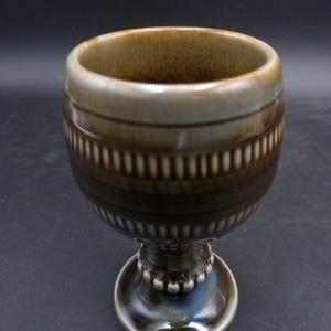 Puede incluir: Un cáliz de cerámica marrón y verde con un diseño estampado. El cáliz tiene una copa ancha y un tallo corto con una base redonda.