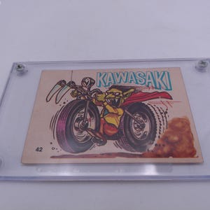 Puede incluir: Una tarjeta de intercambio vintage con un duende verde montando una motocicleta Kawasaki. La tarjeta está enmarcada en una caja de plástico transparente. La tarjeta tiene el número 42 impreso en ella.