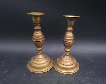 May include: Two antique brass candlesticks with a classic design. Each candlestick has a round base, a detailed stem, and a circular top. The brass has a warm, aged patina, and the candlesticks are set against a dark background.