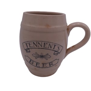 May include: A white ceramic beer mug shaped like a barrel with a blue and white logo that reads "Tennent's Beer".