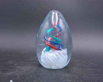 May include: Clear glass paperweight with a swirling design of red, teal, and blue. The interior features a white, cloud-like base. The paperweight has an egg-like shape and is a decorative item.
