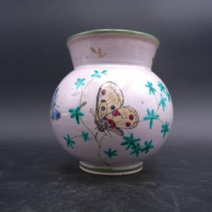 Puede incluir: Un pequeño jarrón redondo de cerámica rosa con un borde verde. El jarrón está decorado con hojas verdes y dos mariposas, una amarilla y otra azul.