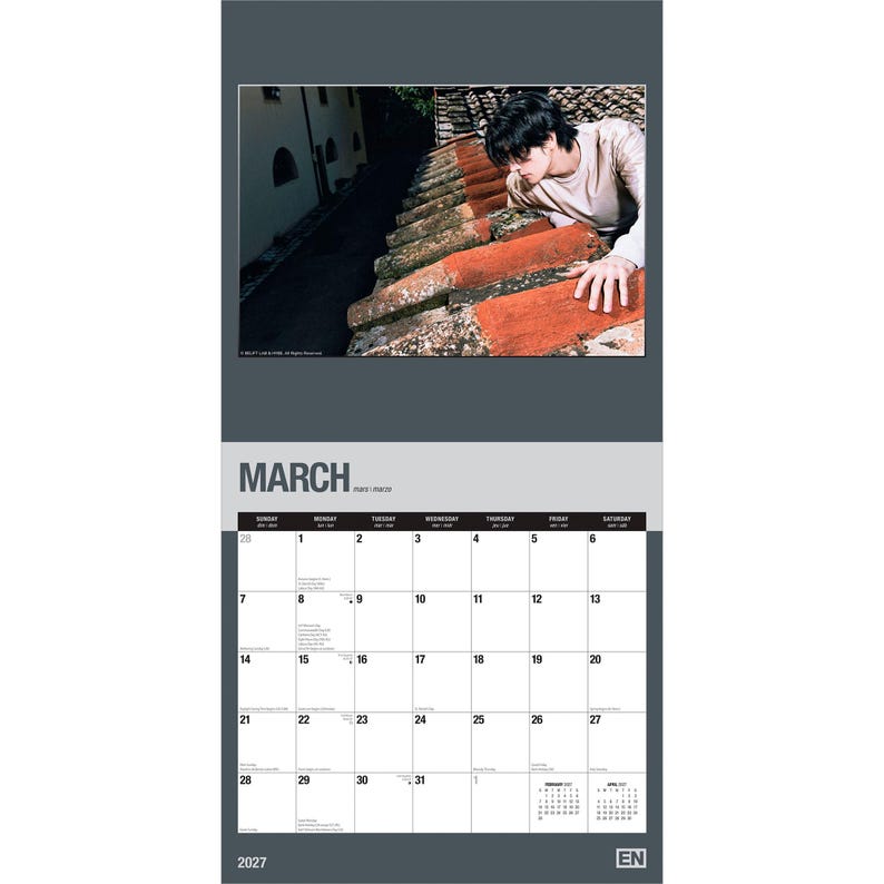 May include: A 2027 wall calendar featuring a photograph of a person in a beige long-sleeve shirt leaning over a weathered terracotta tiled roof. The calendar page displays the month of March with a grid layout, dates, and small text.