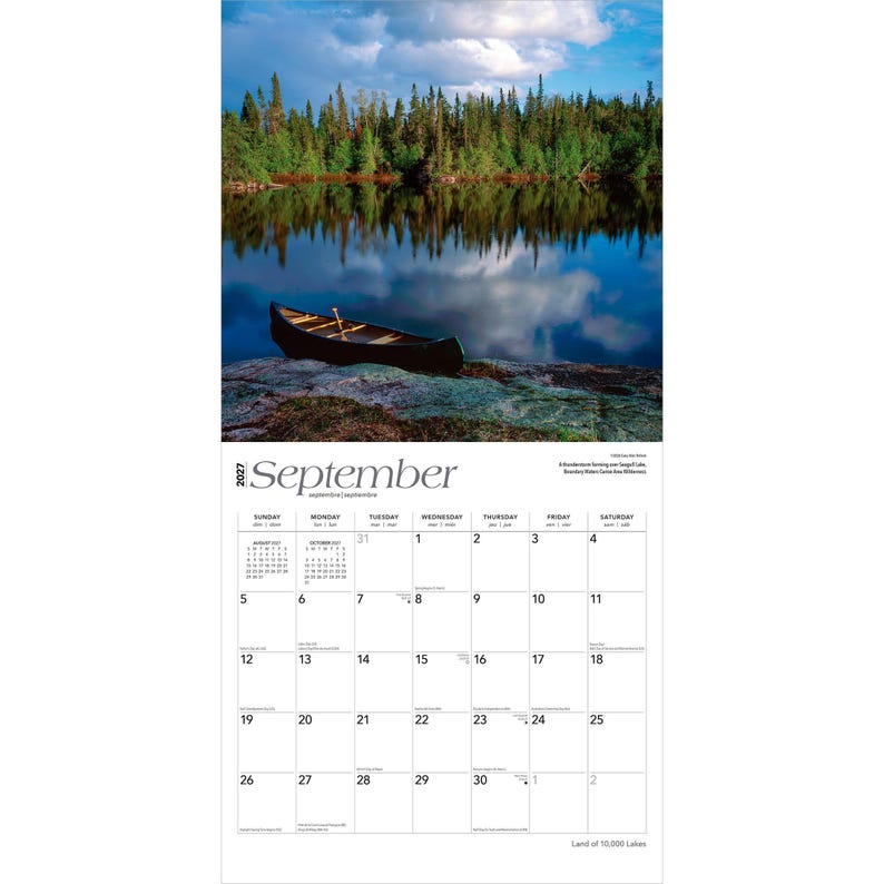May include: A 2027 wall calendar for September featuring a photograph of a dark canoe resting on a rocky shore by a calm lake. The background shows a dense forest reflecting in the water under a cloudy sky. The calendar grid displays dates.