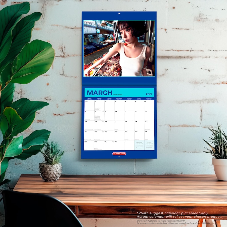 May include: A wall calendar for March 2027 featuring a photograph of a person with short dark hair wearing a white tank top. The calendar has a blue border, a grid layout with numbered days, and the text LE SSERAFIM printed at the bottom center.