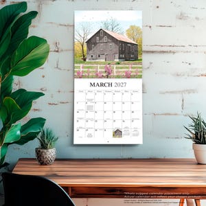 K&ouml;nnte beinhalten: Ein Wandkalender f&uuml;r M&auml;rz 2027 mit einem Foto einer gro&szlig;en, verwitterten braunen Scheune hinter einem wei&szlig;en Holzzaun mit rosa Blumen im Vordergrund. Das Kalenderraster zeigt die Tage des Monats mit deutlichem, schwarzem Text.