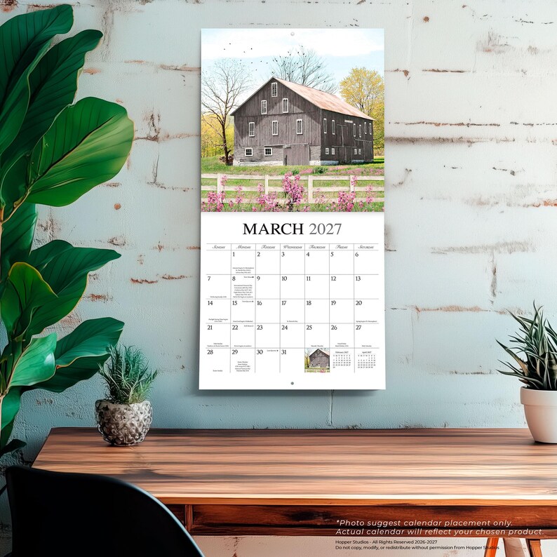 K&ouml;nnte beinhalten: Ein Wandkalender f&uuml;r M&auml;rz 2027 mit einem Foto einer gro&szlig;en, verwitterten braunen Scheune hinter einem wei&szlig;en Holzzaun mit rosa Blumen im Vordergrund. Das Kalenderraster zeigt die Tage des Monats mit deutlichem, schwarzem Text.