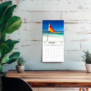 May include: A wall calendar for January 2027 featuring a photograph of a red and yellow flag on a pole at a beach with turquoise water and white sand. The calendar grid displays the days of the month with black text on a white background.