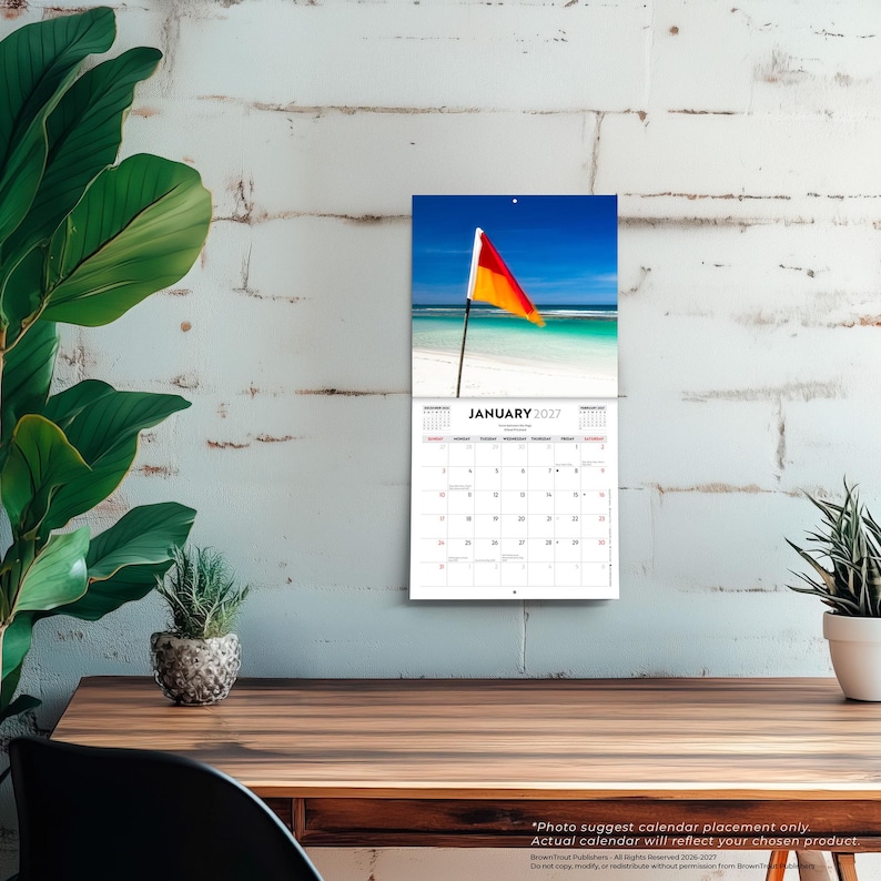 May include: A wall calendar for January 2027 featuring a photograph of a red and yellow flag on a pole at a beach with turquoise water and white sand. The calendar grid displays the days of the month with black text on a white background.