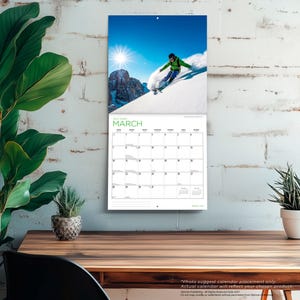 Op de afbeelding: Een wandkalender voor maart 2027 met een foto van een ski&euml;r in een groen jack en blauwe broek die over een besneeuwde berghelling skiet onder een stralende zon. Het kalenderraster onder de afbeelding toont de dagen van de maand overzichtelijk.