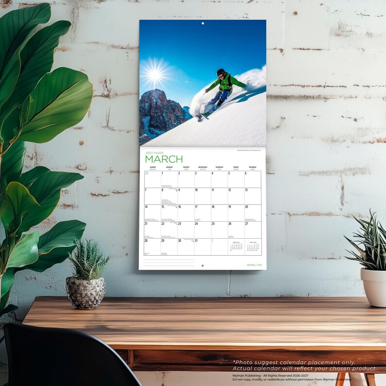 Op de afbeelding: Een wandkalender voor maart 2027 met een foto van een ski&euml;r in een groen jack en blauwe broek die over een besneeuwde berghelling skiet onder een stralende zon. Het kalenderraster onder de afbeelding toont de dagen van de maand overzichtelijk.