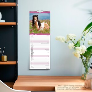 May include: A vertical wall calendar for March 2027 featuring a photograph of a brown and white horse standing in a field of pink flowers. The calendar includes a grid with dates and days of the week listed in black text on a white background.