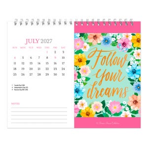 May include: A spiral-bound desk calendar for July 2027. The left page shows the calendar grid with dates and holidays. The right page features a light green background with a floral border and the gold cursive text Follow your dreams.