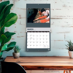 May include: A wall calendar for March 2027 featuring a photograph of a person leaning over a terracotta tiled roof. The calendar grid displays the days of the month with the text MARCH 2027 at the top and the year 2027 at the bottom left corner.
