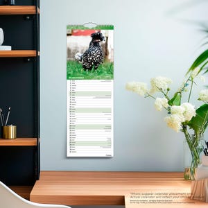 Op de afbeelding: Een verticale wandkalender voor maart 2027 met een foto van een zwart-wit gespikkelde kip met een kuif van veren op de kop, staand in groen gras. Het kalenderraster vermeldt de data 1 tot en met 31 met horizontale lijnen.
