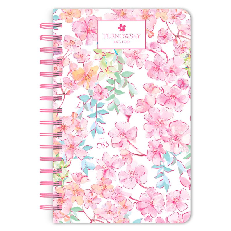 May include: A pink spiral bound notebook with a white background and a floral pattern of pink, yellow, and blue flowers. The notebook has the text "TURNOWSKY EST. 1940" on the front cover.