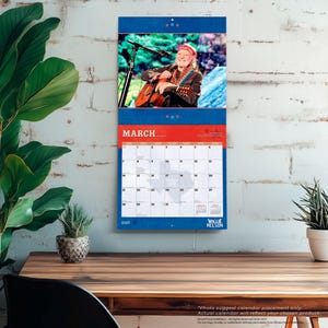 Op de afbeelding: Een wandkalender met een foto van een muzikant die gitaar speelt. De kalender heeft een blauwe en rode rand met de maand maart weergegeven. Het jaar 2027 staat onderaan afgedrukt. De kalender hangt aan een witte bakstenen muur.