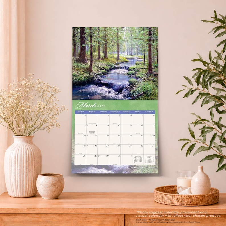 May include: A wall calendar for March 2027 featuring a photograph of a forest stream flowing over rocks. The calendar grid is printed on a light green background with black text for the days of the week and dates, including small calendars for February and April.