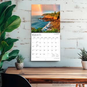 May include: A wall calendar for March 2027 featuring a landscape photograph of a rocky coastline with a rainbow over the ocean. The calendar grid displays the days of the month in black text on a white background with the title March at the top.