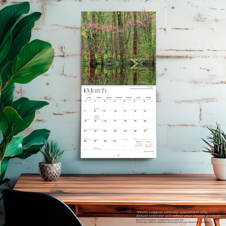 May include: A wall calendar for March 2027 featuring a photograph of a forest with pink flowering trees reflected in a body of water. The calendar grid displays the days of the month with the text March 2027 clearly printed at the top of the page.