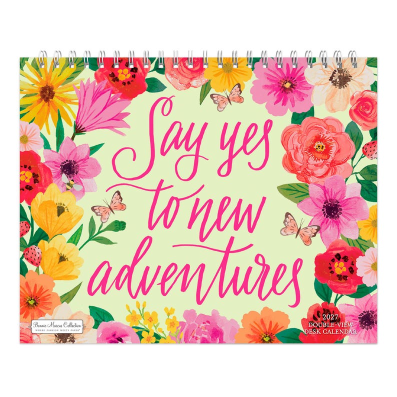 May include: A 2027 desk calendar with a spiral binding at the top. The cover features a light yellow background with a vibrant floral border of pink, red, yellow, and white flowers and butterflies. The text reads: Say yes to new adventures.