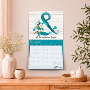 May include: A wall calendar for March 2027 featuring a white wood-plank background with a large teal ampersand symbol decorated with white and teal flowers. The text reads "so the adventure begins." and "LOVE by Fancy" at the bottom.