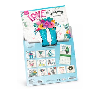 May include: A 2027 wall calendar titled Love the Journey with artwork by Jo Moulton. The cover features bright teal rain boots filled with colourful flowers. The back displays twelve monthly grid pages with various floral and seasonal illustrations.