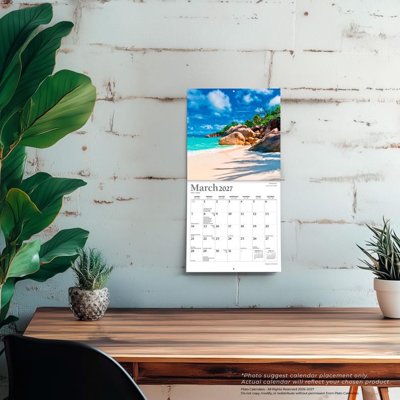 May include: A wall calendar for March 2027 featuring a photograph of a tropical beach with white sand, turquoise water, and large boulders under a blue sky. The calendar grid displays the days of the month with black text on a white background.