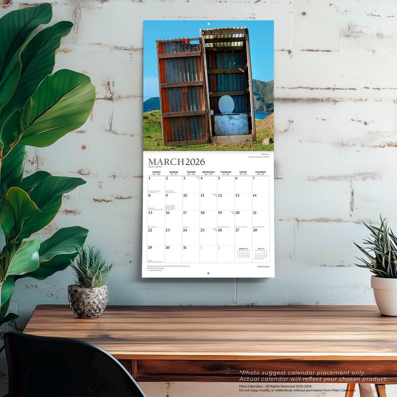 May include: A wall calendar for March 2026 featuring a rustic outhouse with an open door, set against a scenic backdrop. The calendar is displayed on a wooden desk with a plant in a decorative pot. The text "MARCH 2026" is visible.