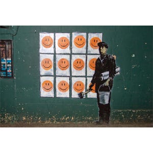 May include: A street art piece featuring a green wall with a grid of orange smiley faces. A silhouetted figure of a painter holding a brush, bucket, and rolled papers stands in front of the wall. The artwork has a political or social commentary theme.