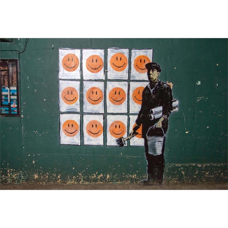 May include: A street art piece featuring a green wall with a grid of orange smiley faces. A silhouetted figure of a painter holding a brush, bucket, and rolled papers stands in front of the wall. The artwork has a political or social commentary theme.