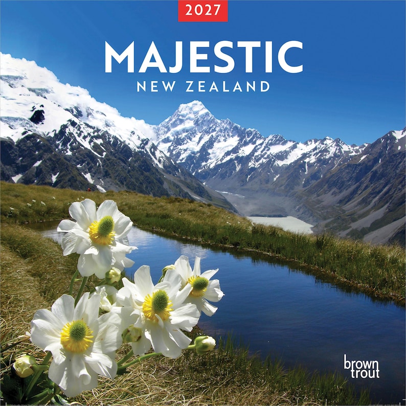 May include: A 2027 wall calendar titled Majestic New Zealand featuring a photograph of white flowers with yellow centers in the foreground, a small stream, and snow-capped mountains under a clear blue sky. The brand name BrownTrout is visible.